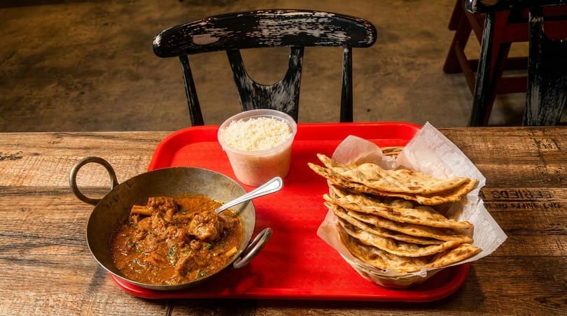 A red tray holds a metal bowl of curry, a container of rice, and a basket of flatbread.