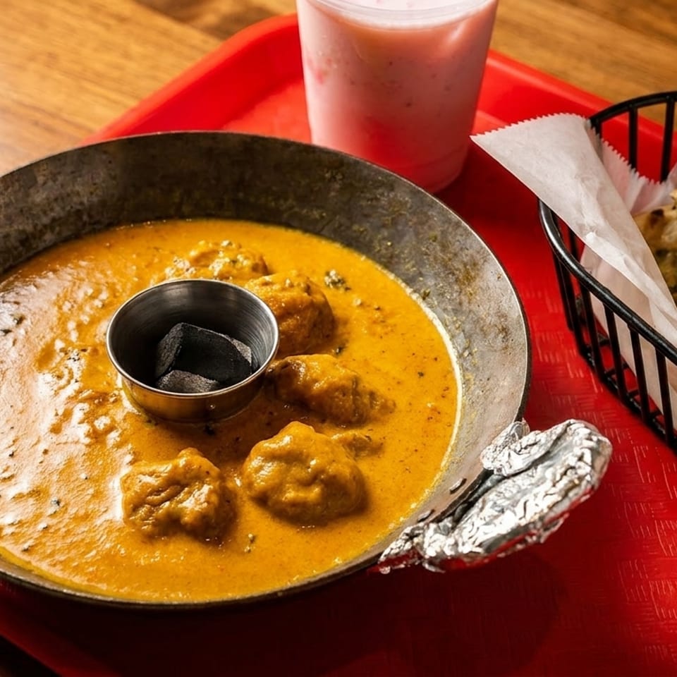 Chicken curry in a metal pan with naan bread in a basket and a pink drink on a red tray.