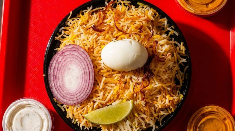 Biryani with a boiled egg, red onion, and lime wedge, served with sauces on a red tray.