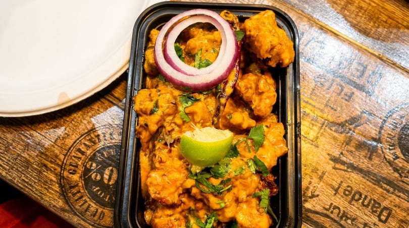 Spicy orange chicken with red onion and lime in a black takeout container on a wooden table.